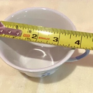 May include: A white teacup with a blue handle and floral design is measured with a yellow tape measure. The cup's diameter is approximately 4 inches. The cup is on a light yellow surface.