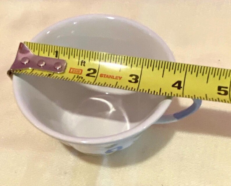 May include: A white teacup with a blue handle and floral design is measured with a yellow tape measure. The cup's diameter is approximately 4 inches. The cup is on a light yellow surface.
