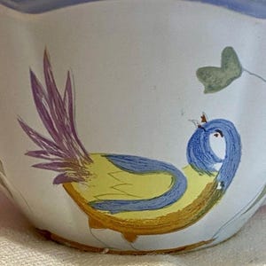 May include: A white ceramic bowl with a hand-painted bird design. The bird features a yellow body, blue head, and purple tail feathers. Green leaves and a blue rim complete the design.