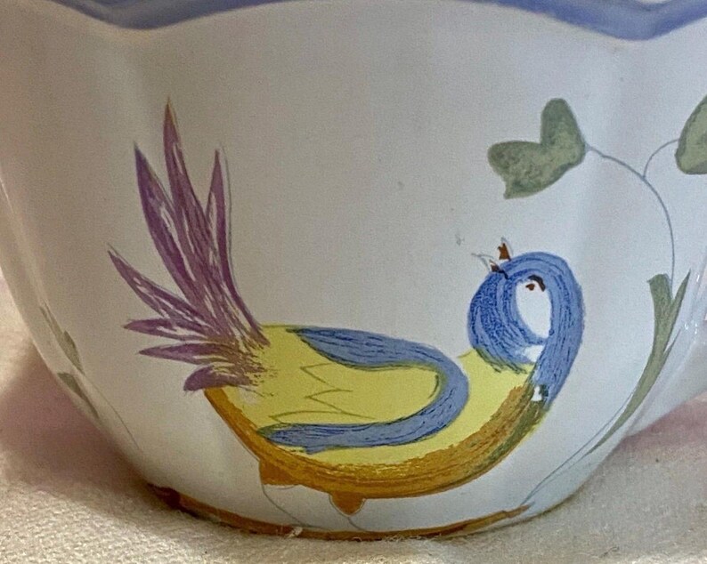 May include: A white ceramic bowl with a hand-painted bird design. The bird features a yellow body, blue head, and purple tail feathers. Green leaves and a blue rim complete the design.