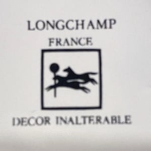 May include: A white background with the words "LONGCHAMP FRANCE" in black text. Below is a black square containing a silhouette of a horse and rider. The words "DECOR INALTERABLE" are below the square.