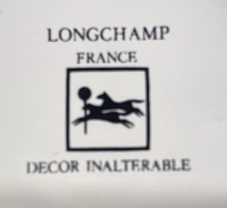 May include: A white background with the words "LONGCHAMP FRANCE" in black text. Below is a black square containing a silhouette of a horse and rider. The words "DECOR INALTERABLE" are below the square.