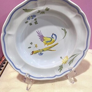 May include: White ceramic plate with a scalloped edge and a blue border. The plate features a hand-painted design of a yellow and purple bird perched on a branch with floral accents. The plate is displayed on a clear acrylic stand.