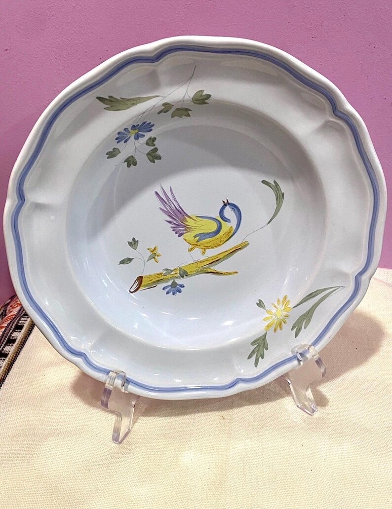 May include: White ceramic plate with a scalloped edge and a blue border. The plate features a hand-painted design of a yellow and purple bird perched on a branch with floral accents. The plate is displayed on a clear acrylic stand.