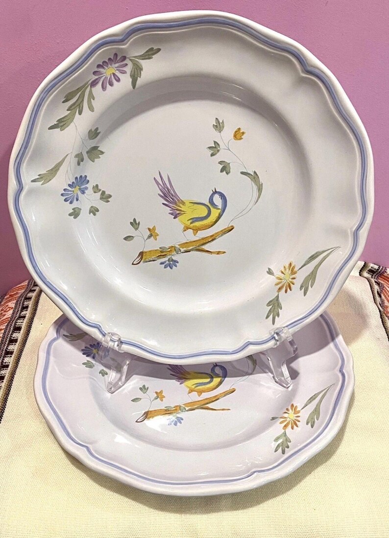May include: Two white ceramic plates with a scalloped edge and a blue border. Each plate features a hand-painted design of a yellow bird perched on a branch, surrounded by flowers and foliage.
