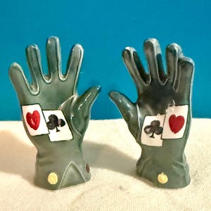 Puede incluir: Un par de saleros y pimenteros de cerámica con forma de mano. Los saleros son de color verde apagado y presentan diseños de palos de cartas: corazones y tréboles. Cada salero tiene una pequeña base redonda amarilla.