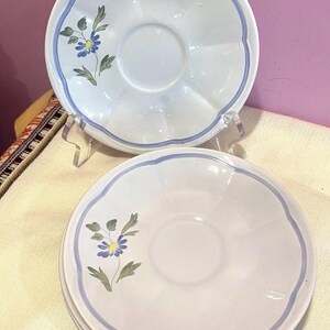 May include: White ceramic saucer plates with a scalloped edge and a blue border. Each plate features a floral design with blue and yellow flowers and green leaves. The plates are stacked and displayed on a clear stand.