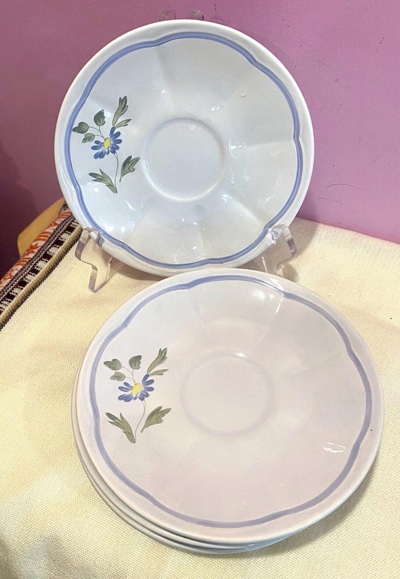 May include: White ceramic saucer plates with a scalloped edge and a blue border. Each plate features a floral design with blue and yellow flowers and green leaves. The plates are stacked and displayed on a clear stand.