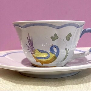 May include: A white ceramic teacup and saucer set. The cup and saucer have a scalloped edge with a blue trim. The cup features a hand-painted bird design in yellow, blue, and purple. The handle is blue.