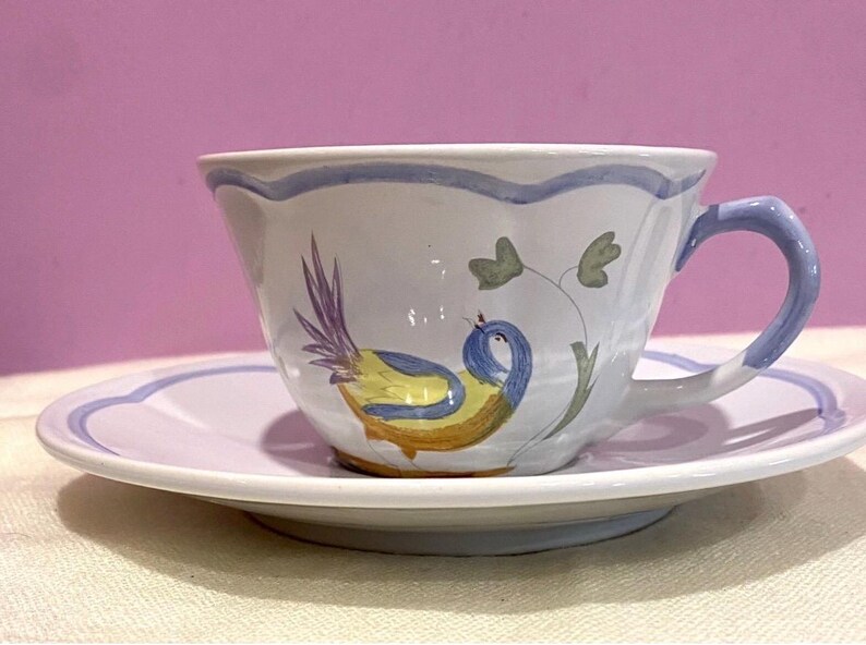 May include: A white ceramic teacup and saucer set. The cup and saucer have a scalloped edge with a blue trim. The cup features a hand-painted bird design in yellow, blue, and purple. The handle is blue.