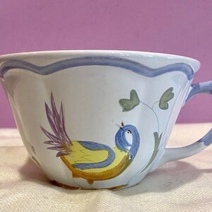 May include: White ceramic teacup with a blue handle and rim. The cup features a hand-painted design of a yellow and blue bird with purple tail feathers and green leaves. The cup is on a white surface.