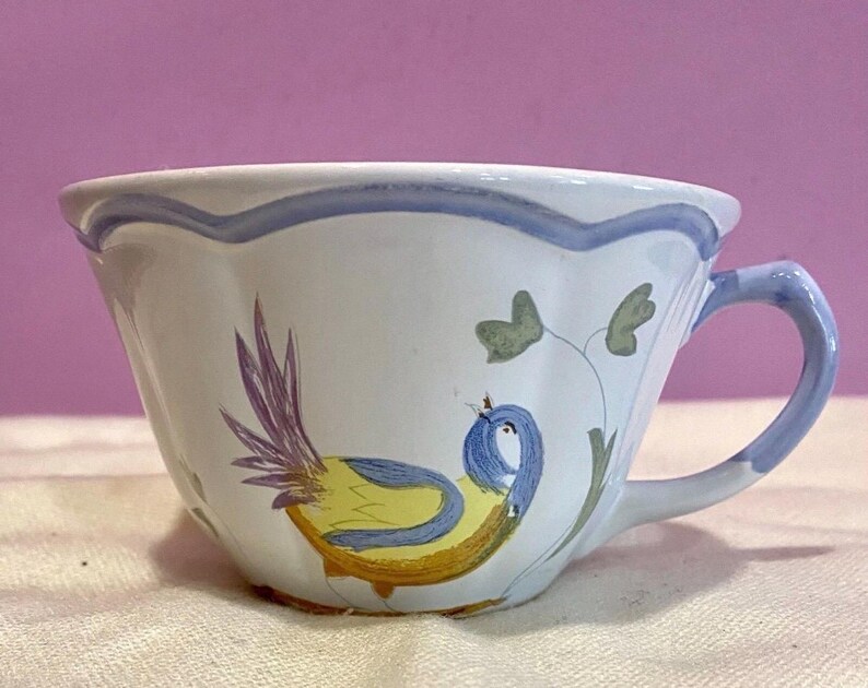 May include: White ceramic teacup with a blue handle and rim. The cup features a hand-painted design of a yellow and blue bird with purple tail feathers and green leaves. The cup is on a white surface.