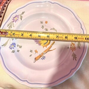Pu&ograve; includere: Un piatto in ceramica bianca con un motivo floreale blu e viola e un uccello giallo con un ramo marrone. Il piatto ha un bordo a festoni e misura 23 cm di diametro.