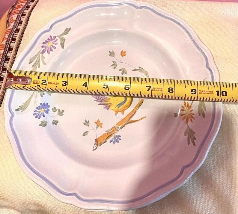 Pu&ograve; includere: Un piatto in ceramica bianca con un motivo floreale blu e viola e un uccello giallo con un ramo marrone. Il piatto ha un bordo a festoni e misura 23 cm di diametro.