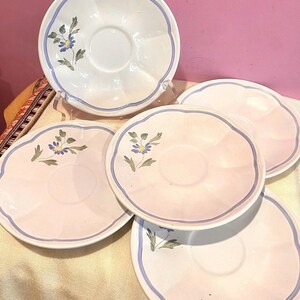 May include: A collection of white and pink saucers with a blue floral design. Each saucer has a scalloped edge and a blue border. The floral design features blue flowers and green leaves. The saucers are arranged on a light-colored surface.