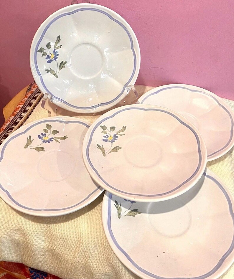 May include: A collection of white and pink saucers with a blue floral design. Each saucer has a scalloped edge and a blue border. The floral design features blue flowers and green leaves. The saucers are arranged on a light-colored surface.