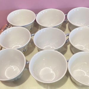 May include: Nine white ceramic teacups with light blue handles and floral designs. The cups are arranged in three rows of three, showcasing their clean, simple design. The cups are on a patterned surface.