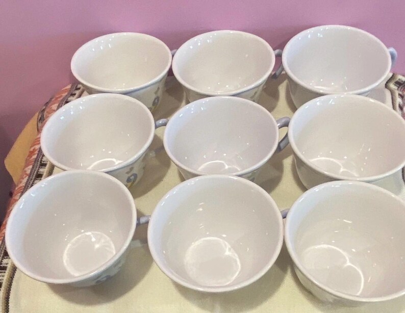 May include: Nine white ceramic teacups with light blue handles and floral designs. The cups are arranged in three rows of three, showcasing their clean, simple design. The cups are on a patterned surface.