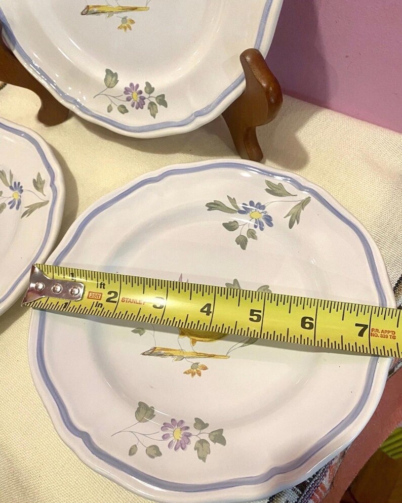 May include: White ceramic plates with a blue rim and floral design. The plates are approximately 7 inches in diameter. The plates have a scalloped edge and are decorated with purple and yellow flowers.