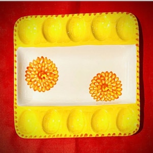 Pode incluir: Um prato de ovos de cerâmica branca com uma borda amarela e seis porta-ovos amarelos. O prato tem dois desenhos florais laranja e amarelo.