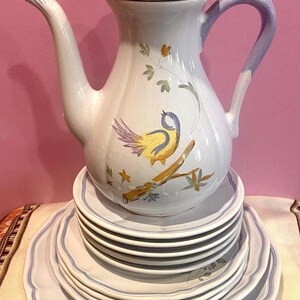 May include: A white ceramic teapot with a lid, a curved handle, and a spout, decorated with a yellow and blue bird on a branch. The teapot sits atop a stack of matching plates with blue trim and floral designs.