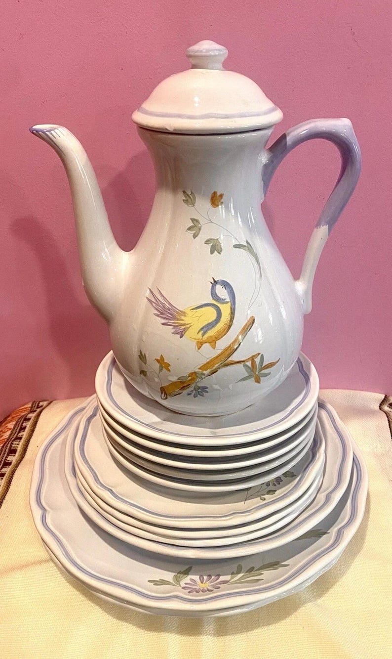 May include: A white ceramic teapot with a lid, a curved handle, and a spout, decorated with a yellow and blue bird on a branch. The teapot sits atop a stack of matching plates with blue trim and floral designs.