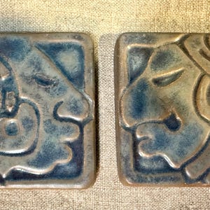 May include: Two square ceramic tiles featuring embossed profiles of figures in a blue and brown glaze. Each tile displays a stylized, relief-carved face with intricate details, finished with a glossy, mottled blue and earthy brown color palette.