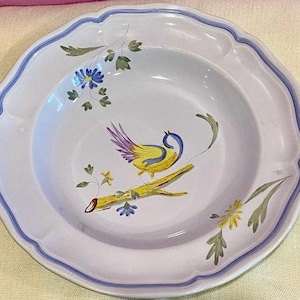 Pu&ograve; includere: Una ciotola per la zuppa in ceramica bianca con un bordo blu e un motivo floreale. La ciotola presenta un uccello giallo con ali viola appollaiato su un ramo con foglie verdi e fiori gialli.