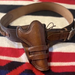 May include: A brown leather gun holster and belt set. The holster is attached to a wide belt with bullet loops. The belt has a buckle and is designed to be worn around the waist. The set is made of brown leather.