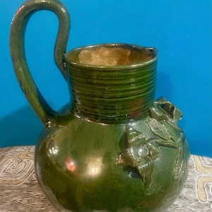 Op de afbeelding: Een groene keramische kan met een grote handgreep en een afgeronde body. De kan heeft een glanzende afwerking en is versierd met reliëf bloemmotieven. De rand en de handgreep zijn een donkerdere tint groen.