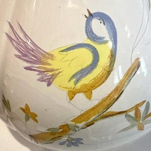 Pu&ograve; includere: Un'anfora in ceramica bianca con un uccello giallo appollaiato su un ramo. L'uccello ha le ali blu e una coda rosa. L'anfora &egrave; decorata con fiori gialli e verdi.