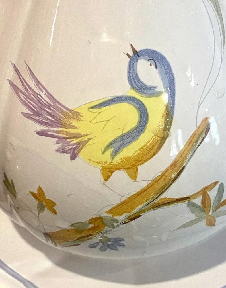 Pu&ograve; includere: Un'anfora in ceramica bianca con un uccello giallo appollaiato su un ramo. L'uccello ha le ali blu e una coda rosa. L'anfora &egrave; decorata con fiori gialli e verdi.