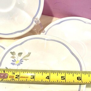 May include: Several white ceramic saucers with a light blue scalloped edge and a blue floral design. A yellow tape measure shows the saucers are approximately 6 inches in diameter.
