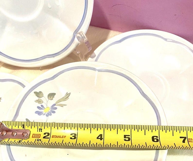 May include: Several white ceramic saucers with a light blue scalloped edge and a blue floral design. A yellow tape measure shows the saucers are approximately 6 inches in diameter.