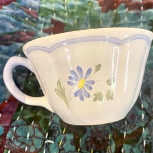 May include: A white ceramic teacup with a light blue handle and rim. The cup features a painted blue daisy with a yellow center and green leaves. The cup is on a patterned fabric background.