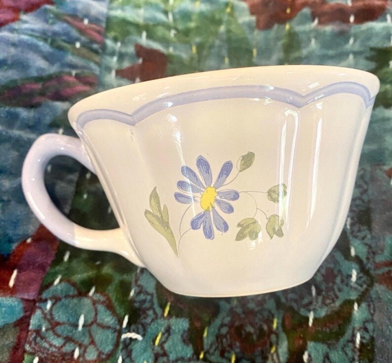 May include: A white ceramic teacup with a light blue handle and rim. The cup features a painted blue daisy with a yellow center and green leaves. The cup is on a patterned fabric background.