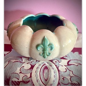 May include: A white ceramic planter with a green and blue fleur-de-lis design. The planter has a scalloped edge and is sitting on a patterned fabric.