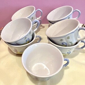May include: A set of white ceramic teacups with blue handles and rims. Each cup is decorated with a floral and bird design in shades of blue and yellow. The cups are stacked and arranged on a light-colored surface.