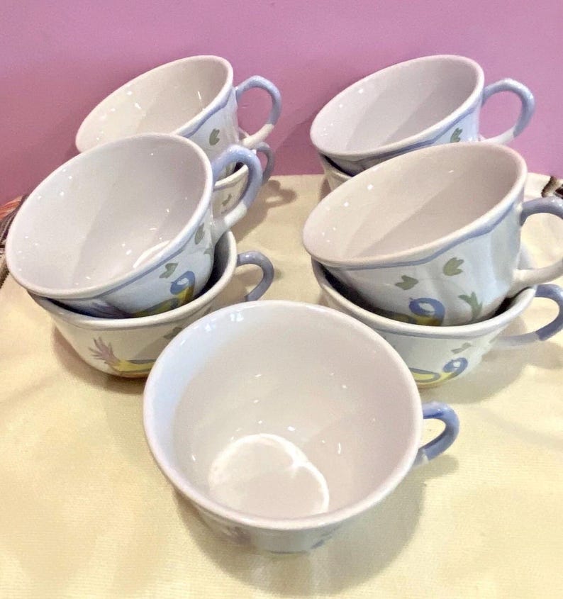 May include: A set of white ceramic teacups with blue handles and rims. Each cup is decorated with a floral and bird design in shades of blue and yellow. The cups are stacked and arranged on a light-colored surface.