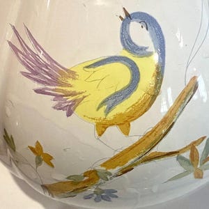 May include: A white ceramic pitcher with a hand-painted design of a yellow bird with blue and purple accents perched on a branch. The bird is singing, and the branch has green leaves and small blue flowers.