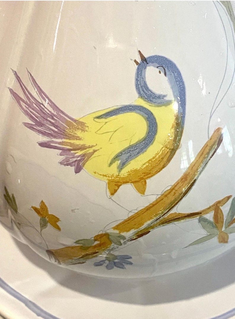 May include: A white ceramic pitcher with a hand-painted design of a yellow bird with blue and purple accents perched on a branch. The bird is singing, and the branch has green leaves and small blue flowers.