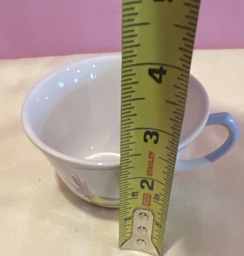 May include: A white teacup with a light blue handle and a floral design. The cup is approximately 3 inches tall, as measured by a yellow tape measure.