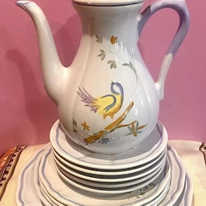 Może przedstawiać: Biały ceramiczny czajnik z niebieskim uchwytem i wzorem żółtego ptaka. Czajnik stoi na stosie białych ceramicznych talerzy z niebieską obwódką.