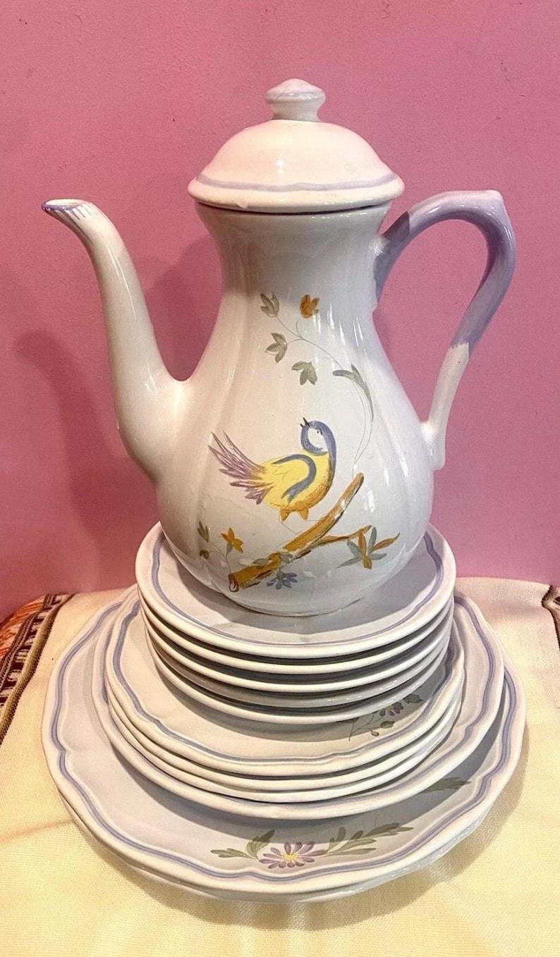 Pu&ograve; includere: Una teiera in ceramica bianca con manico blu e un design di uccello giallo. La teiera &egrave; appoggiata su una pila di piatti in ceramica bianca con bordo blu.
