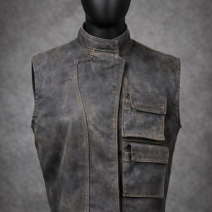 May include: A weathered, dark gray vest with a stand-up collar and three front pockets. The vest has a slightly distressed appearance, with visible stitching and a worn texture. The vest is displayed on a black mannequin.