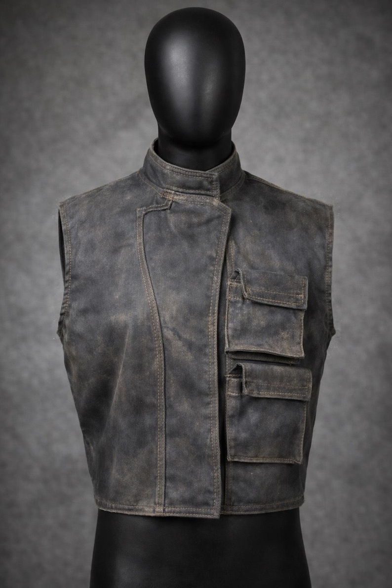 May include: A weathered, dark gray vest with a stand-up collar and three front pockets. The vest has a slightly distressed appearance, with visible stitching and a worn texture. The vest is displayed on a black mannequin.