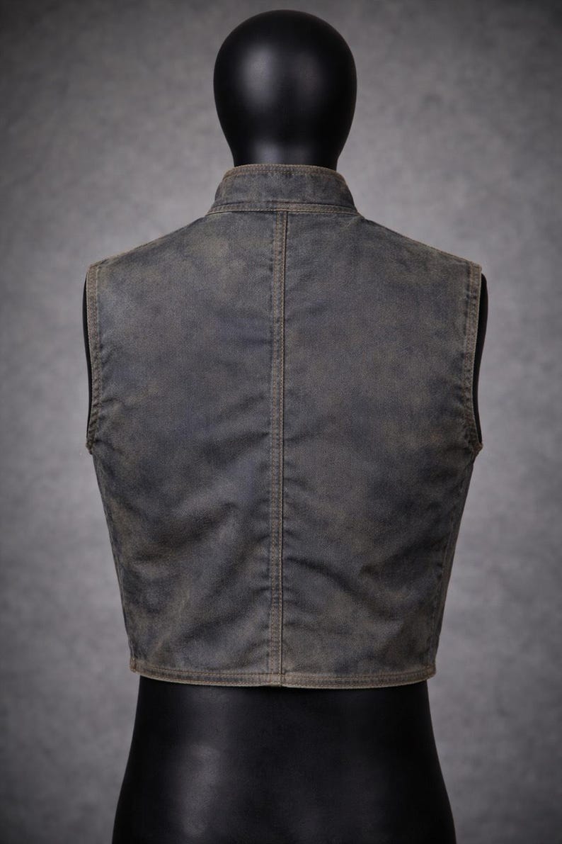 May include: A weathered-look vest in a muted gray-brown hue, displayed on a mannequin. The vest features a stand-up collar and a vertical seam down the back. The fabric has a slightly distressed appearance.