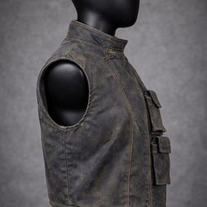 May include: A weathered, gray vest with a stand-up collar and multiple front pockets. The vest has a distressed appearance, suggesting a vintage or rugged style. The sleeveless design and tailored fit are visible.