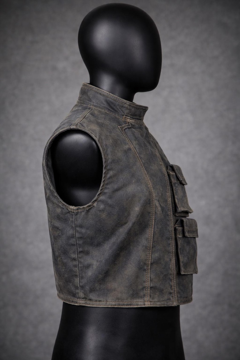 May include: A weathered, gray vest with a stand-up collar and multiple front pockets. The vest has a distressed appearance, suggesting a vintage or rugged style. The sleeveless design and tailored fit are visible.
