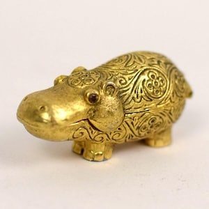 May include: A gold-colored, decorative hippo figurine with intricate floral patterns. The hippo has small, dark red eyes and is posed in a standing position. The figurine is likely made of metal and has a detailed, embossed design.